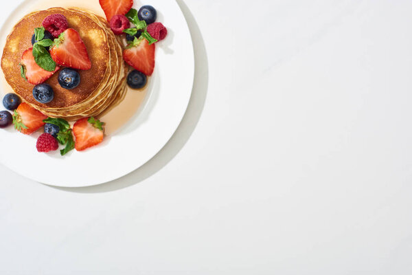 top view of delicious pancakes with honey, blueberries and strawberries on plate on marble white surface