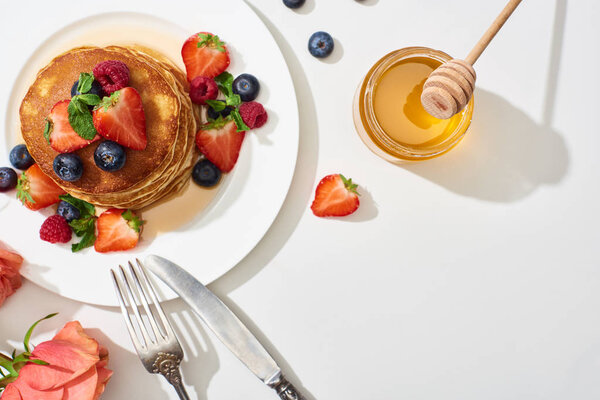 top view of delicious pancakes with honey, blueberries and strawberries on plate near cutlery and rose on marble white surface