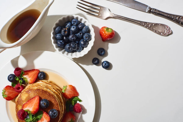top view of delicious pancakes with blueberries and strawberries on plate near cutlery and maple syrup in gravy boat on marble white surface