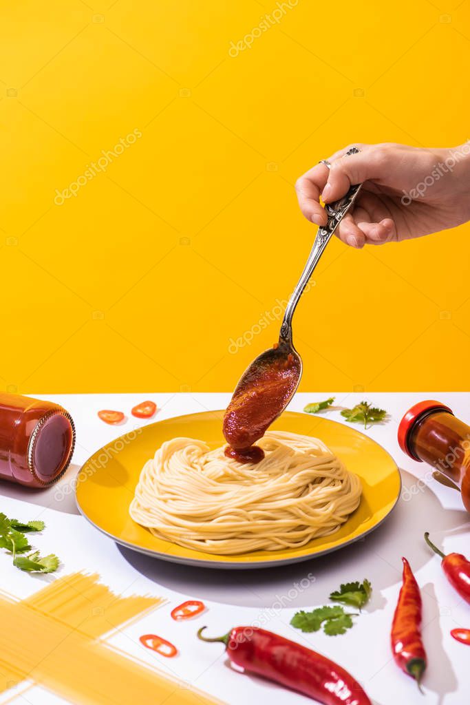 Cropped view of woman pouring ketchup on spaghetti beside chili peppers and cilantro on white surface isolated on yellow