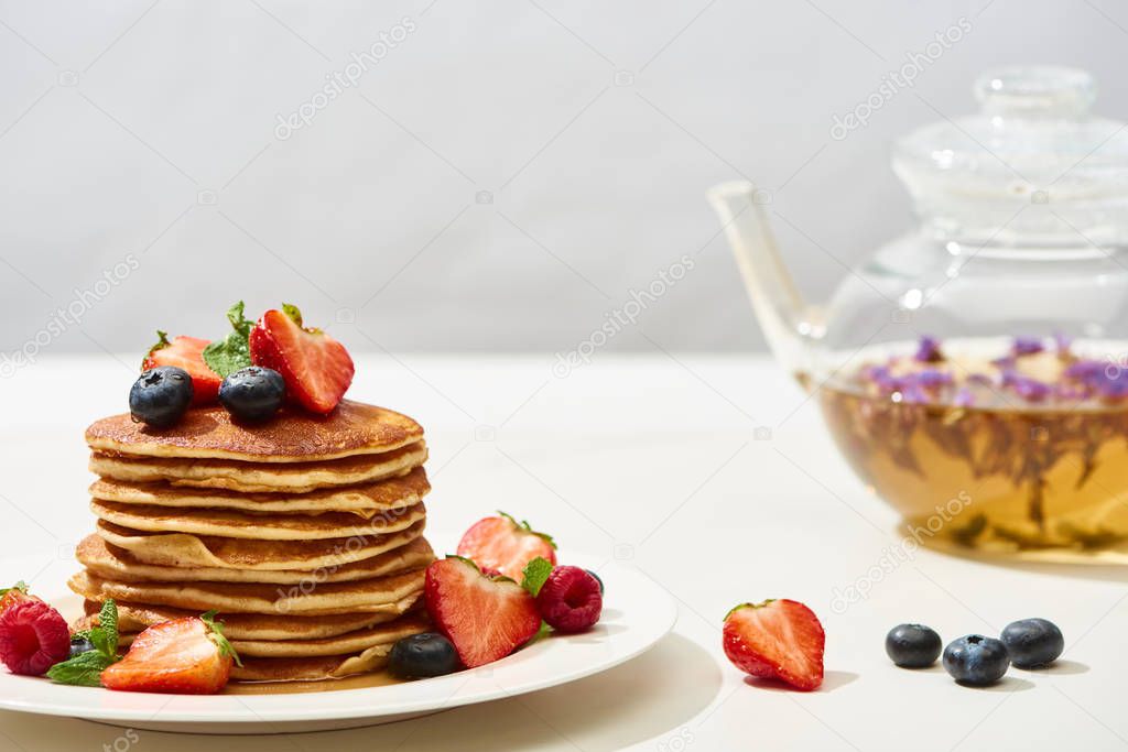 selective focus of delicious pancakes with blueberries and strawberries near herbal tea on white surface isolated on grey