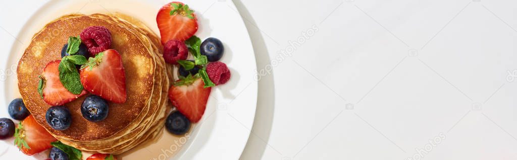 top view of delicious pancakes with honey, blueberries and strawberries on plate on marble white surface, panoramic shot