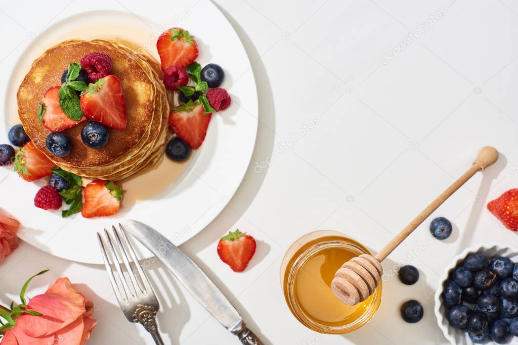 top view of delicious pancakes with honey, blueberries and strawberries on plate near cutlery and rose on marble white surface