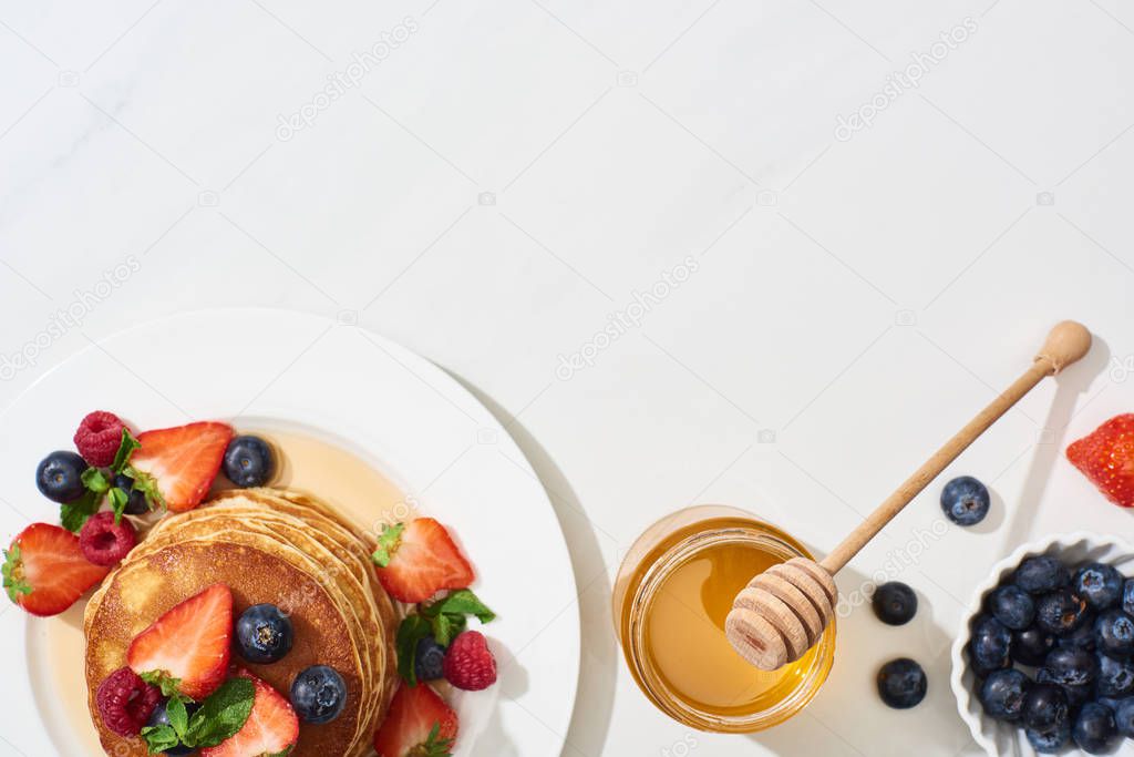 top view of delicious pancakes with honey, blueberries and strawberries on plate on marble white surface