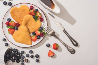 top view of delicious heart shaped pancakes with berries near maple syrup and cutlery on white background clipart