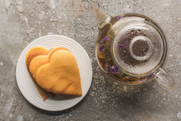 top view of heart shaped pancakes near herbal tea in teapot on grey concrete surface