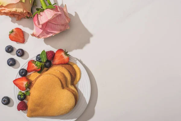白い背景にバラの近くに果実があるおいしいハート型のパンケーキのトップビュー — ストック写真