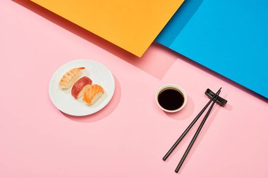 fresh nigiri with salmon, tuna and shrimp near soy sauce and chopsticks on blue, pink, orange surface clipart