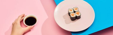 cropped view of woman holding soy sauce near fresh maki with salmon on blue, pink background, panoramic shot clipart