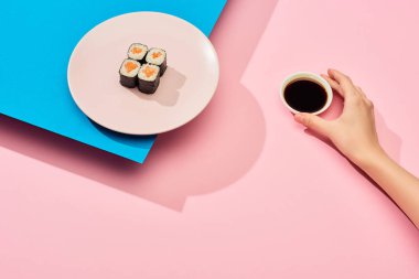 Taze maki yanında soya sosu tutan mavi, pembe arka planda somon balığı tutan kadın görüntüsü.