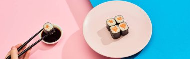 cropped view of woman eating fresh maki with salmon near soy sauce on blue, pink background, panoramic shot clipart