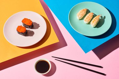 Karidesli taze nigiri ve soya sosunun yanında kırmızı havyar ve mavi, pembe, turuncu arka planda çubuklar.