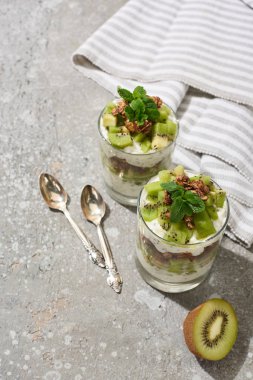 Gri beton yüzeyinde kivi ve yoğurt ve çizgili peçete ve kaşıklarla taze granola.