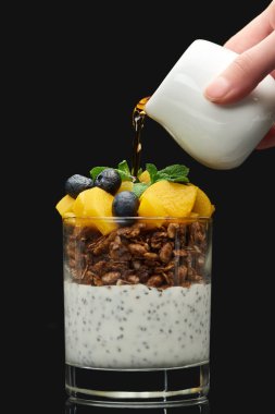 cropped view of woman adding syrup to fresh granola with canned peach, blueberries and chia seeds isolated on black clipart