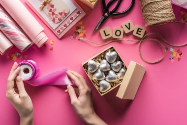 partial view of woman holding spool of ribbon near gift box with silver hearts and valentines handiwork supplies on pink background clipart