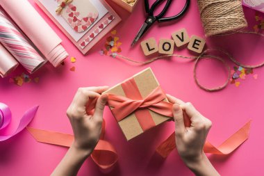 partial view of woman decorating gift box with ribbon near valentines handiwork supplies on pink background clipart