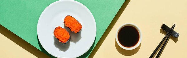 top view of fresh nigiri with red caviar near soy sauce and chopsticks on green and yellow surface, panoramic shot