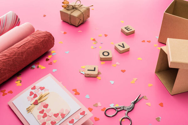 валентинки конфетти, ножницы, оберточная бумага, подарочные коробки, поздравительная открытка и любовная надпись на деревянных кубиках на розовом фоне
