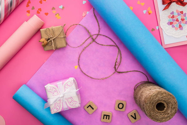 верхний вид украшения валентинки, оберточная бумага, шпагат, подарочные коробки, поздравительная открытка и любовная надпись на деревянных кубиках на розовом фоне
