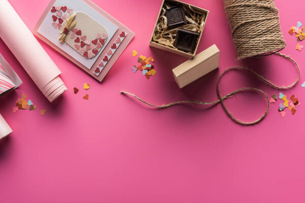 top view of valentines decoration, wrapping paper, twine, gift box with chocolate, greeting card on pink background