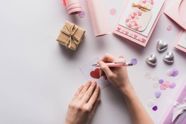 cropped view of woman writing on card near valentines decoration, gifts, hearts and wrapping paper on white background clipart