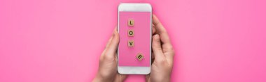 Pembe arka planda aşk harfi yazılı küplerin olduğu akıllı telefonu tutan kadının kısmi görüntüsü, panoramik çekim