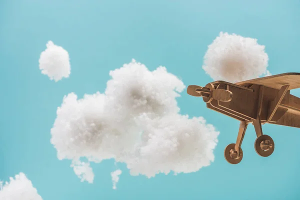 青い上に隔離された綿ウールで作られた白いふわふわの雲の間を飛ぶ木製のおもちゃの飛行機 — ストック写真