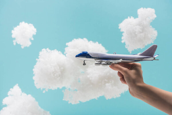 cropped view of woman playing with toy plane among white fluffy clouds made of cotton wool isolated on blue