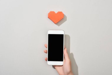 Gri arkaplanda kalp şeklinde kağıt ile boş ekranlı akıllı telefonu tutan kızın üst görünümü