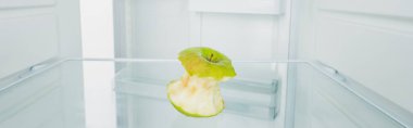 Panoramic shot of gnawed green apple on shelf of refrigerator with open door isolated on white clipart