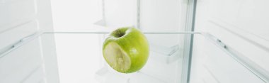 Panoramic shot of bitten off green apple on shelf of refrigerator with open door isolated on white clipart