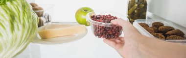 Buzdolabından üzümlü üzümle plastik tepsi alan beyaz panoramik bir karede izole edilmiş bir kadının görüntüsü.