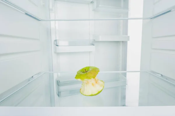 Gnawed green apple on shelf of refrigerator with open door isolated on white