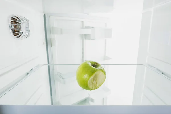 Bitten off green apple on shelf of refrigerator with open door isolated on white