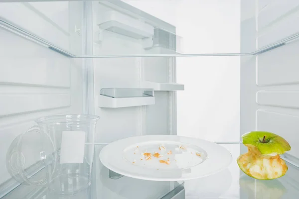 Plastic cup with bitten apple and crumbs on plate in fridge with open door isolated on white