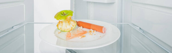 Panoramic shot of bitten apple with sausage and crumbs on plate in fridge with open door isolated on white