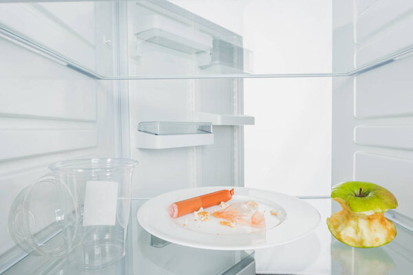Plate with scraps, bitten apple and plastic cup in fridge with open door isolated on white