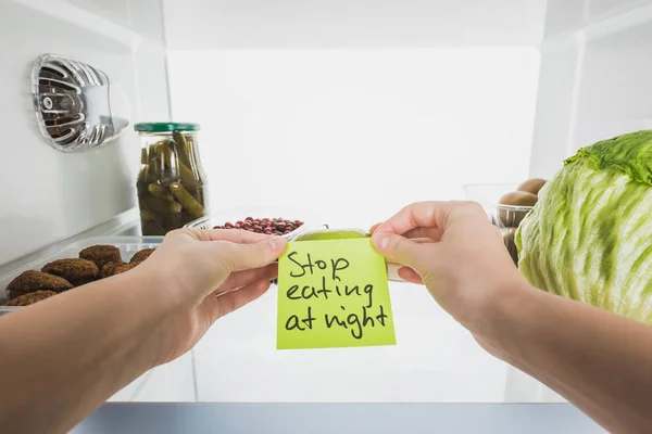 Cropped view of woman holding card with stop eating at night lettering with food in fridge isolated on white