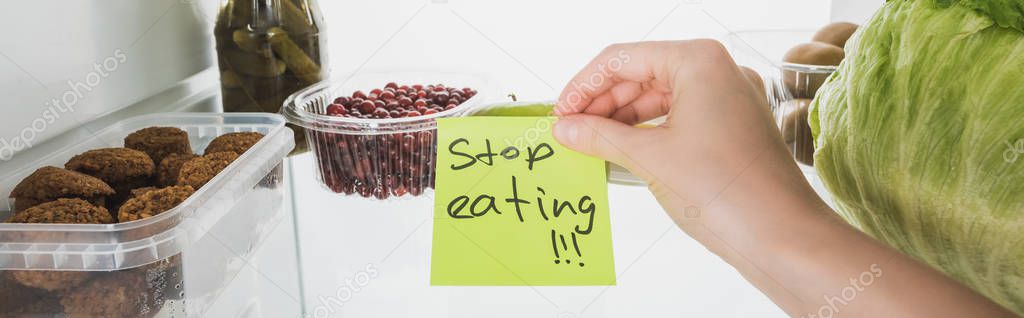 Panoramic shot of food in fridge and woman holding card with stop eating lettering isolated on white