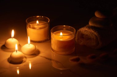 burning candles in glass glowing in dark near rolled towel on marble surface clipart