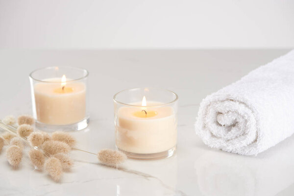 fluffy bunny tail grass near burning white candles in glass and rolled towel on marble white surface