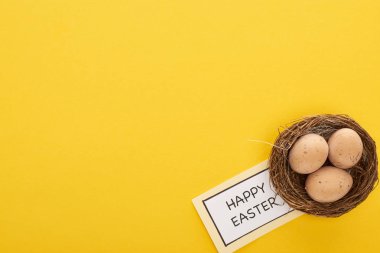 Sarı arka planda tavuk yumurtalarının yanında mutlu Paskalya harfleri olan tebrik kartının üst görüntüsü.