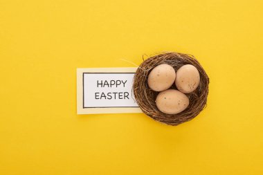 Sarı arka planda tavuk yumurtalarının yanında mutlu Paskalya harfleri olan tebrik kartının üst görüntüsü.