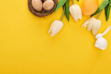 Yuvadaki lalelerin ve tavuk yumurtalarının üst manzarası. Renkli sarı arka planda Paskalya tavşanı.