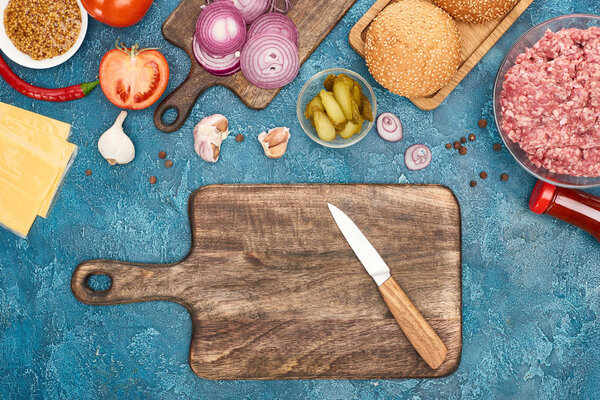 вид сверху на свежие ингредиенты бургера и деревянную доску с ножом на голубой текстурированной поверхности
