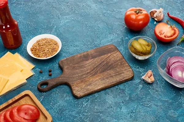 fresh burger ingredients around wooden cutting board on blue textured surface