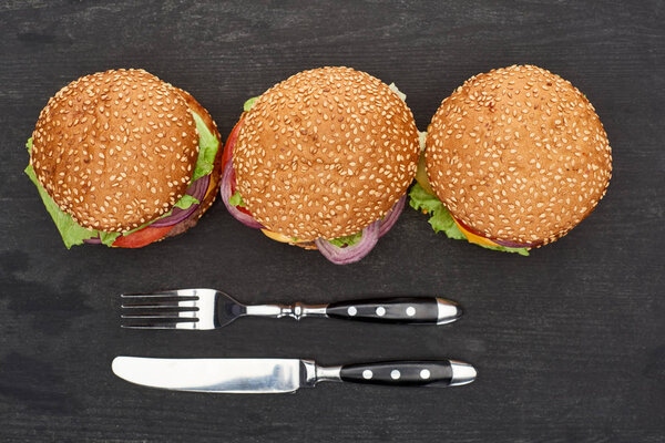 top view of delicious fresh burgers on black wooden table with knife and fork