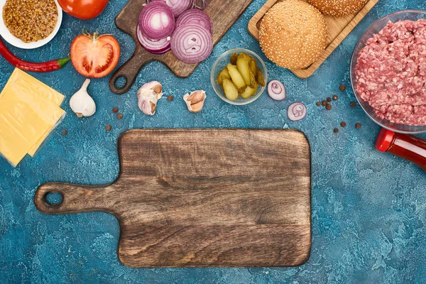 top view of fresh burger ingredients near empty cutting board on blue textured surface