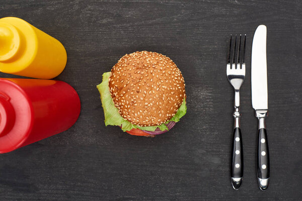 top view of delicious fresh burger near cutlery, ketchup and mustard on black wooden table