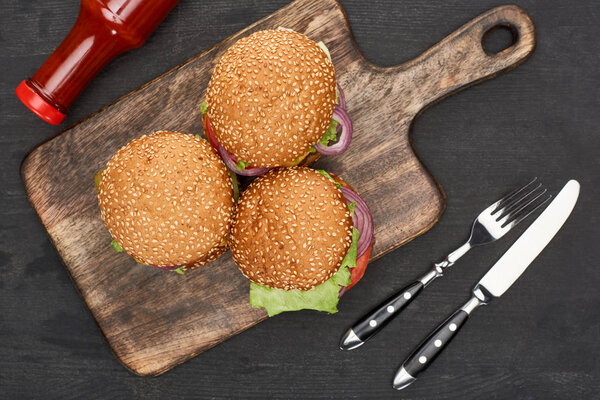 top view of delicious fresh meat burgers on wooden board near cutlery and ketchup